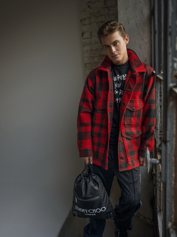 Person wearing a red and black plaid jacket holding a black bag with 'Jimmy Choo' branding.